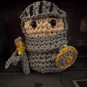 May include: A crocheted knight doll with a gray helmet and armor, a beige face, and black eyes. The knight holds a gold and gray sword and shield. The doll is handmade and has a whimsical design.