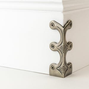 May include: A metallic corner protector with an antique brass finish attached to the corner of a white wooden surface. The hardware features a vertical design with three distinct sections, each secured by small screws, and includes textured diamond patterns.
