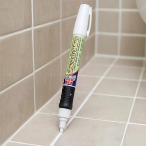 May include: A white grout and tile marker pen with a black grip section. The label features the text Grout & Tile Marker and states it covers stubborn grout stains instantly. The pen has a white cap and a white tip applying white paint.