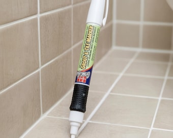 May include: A white grout and tile marker pen with a black grip section. The label features the text Grout & Tile Marker and states it covers stubborn grout stains instantly. The pen has a white cap and a white tip applying white paint.