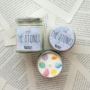 Könnte beinhalten: Drei Vanille-Duftkerzen mit dem Text "THE STONES" auf den Etiketten. Eine befindet sich in einem Glas, zwei in Metalldosen. Die Kerzen sind von Textseiten umgeben. Die Kerze in der Dose hat bunte Kristalle.