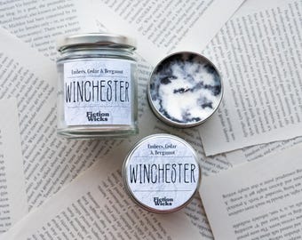 Op de afbeelding: Twee kaarsen in glazen potten met zilveren deksels. De kaarsen zijn gelabeld "Winchester" en hebben verschillende geuren: "Embers, Cedar & Bergamot" en "Embers, Cedar & Bergamot". De kaarsen staan op een achtergrond van witte pagina's.