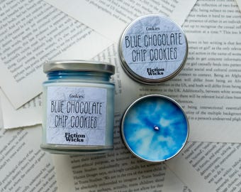 May include: A blue and white candle in a glass jar with a silver lid. The label reads "Cookies Blue Chocolate Chip Cookies" and "Fiction Wicks".