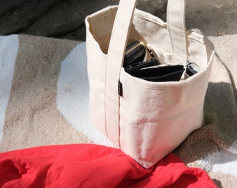 Peut inclure: Un sac fourre-tout en toile de couleur crème avec une poignée, contenant un portefeuille noir et d'autres articles. Une chemise rouge est posée à côté du sac sur une couverture à rayures. La scène est éclairée par la lumière naturelle.