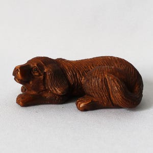May include: A small, intricately carved wooden dog figurine in a resting pose. The dog is a rich brown color, with detailed carving that highlights the fur and facial features. The figurine is lying down, with its paws tucked beneath it.