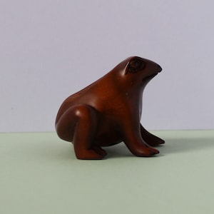 May include: A wooden frog figurine with a brown finish. The frog is sitting on its hind legs with its front legs tucked in. The frog is looking to the right.