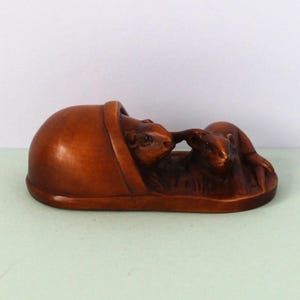May include: A brown carved wooden figurine depicting two small rodents, one resting inside a smooth, rounded shell-like structure and the other positioned beside it on a flat base, showcasing detailed textures and a polished finish throughout.