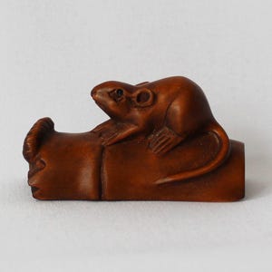 May include: A small, intricately carved wooden sculpture of a rat resting on a log. The rat is brown with detailed features, and the log is a darker shade of brown. The sculpture is set against a plain white background.