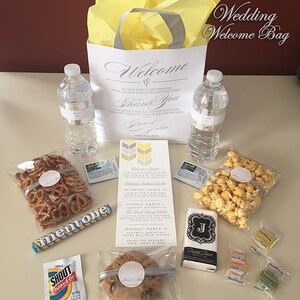 May include: A white wedding welcome bag with a yellow tissue paper lining. The bag contains a variety of items, including water bottles, pretzels, popcorn, a cookie, a hand soap, a packet of tissues, and a few small candies. The bag also includes a wedding invitation card with the couple's names and wedding date.