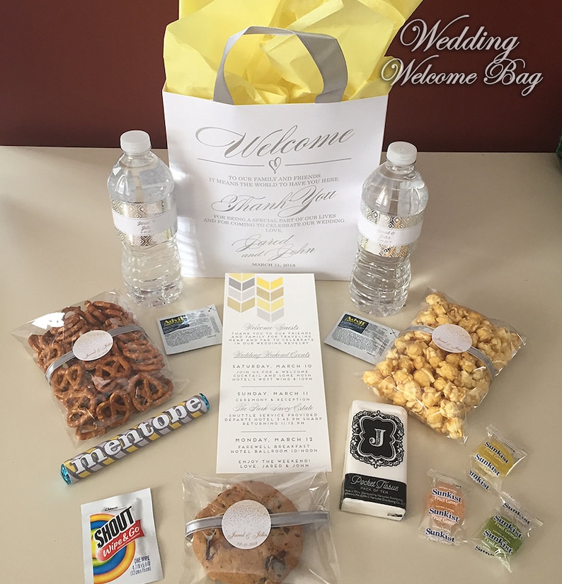 May include: A white wedding welcome bag with a yellow tissue paper lining. The bag contains a variety of items, including water bottles, pretzels, popcorn, a cookie, a hand soap, a packet of tissues, and a few small candies. The bag also includes a wedding invitation card with the couple's names and wedding date.