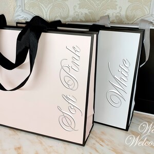 May include: Three wedding welcome bags in soft pink, white, and black with matching ribbon handles. The bags are labeled with the color of the bag in a silver script font. The text at the bottom of the image reads "Wedding Welcome Bag".