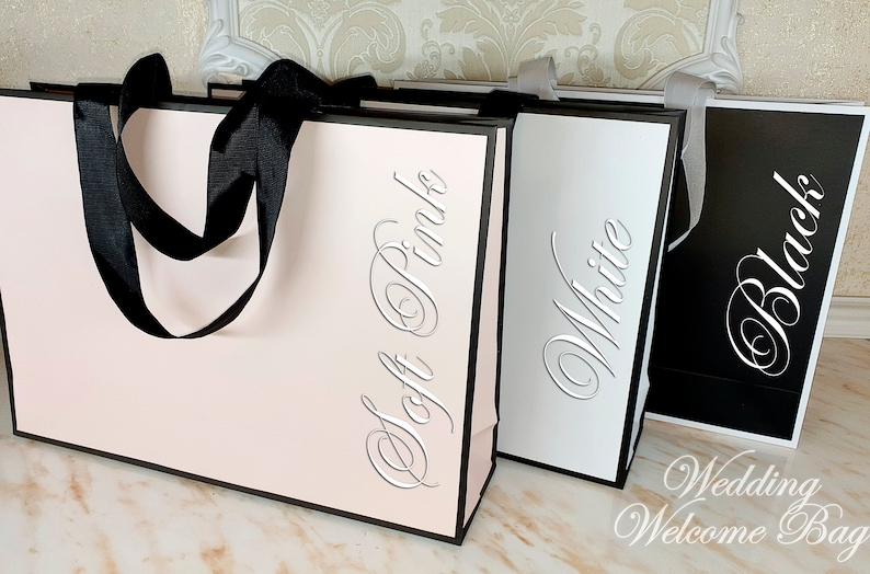 May include: Three wedding welcome bags in soft pink, white, and black with matching ribbon handles. The bags are labeled with the color of the bag in a silver script font. The text at the bottom of the image reads "Wedding Welcome Bag".