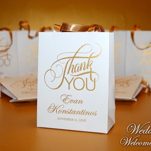 Op de afbeelding: Witte bruiloft-cadeauzakje met gouden lintjes en gouden opschrift "Bedankt" en "Evan Konstantinos" met de datum "11 november 2018".