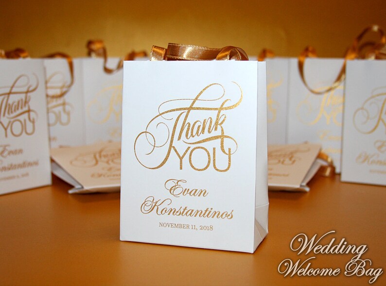 Op de afbeelding: Witte bruiloft-cadeauzakje met gouden lintjes en gouden opschrift "Bedankt" en "Evan Konstantinos" met de datum "11 november 2018".