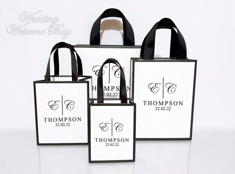 May include: A collection of white gift bags with black trim and handles. Each bag features a personalized design with initials, the last name "THOMPSON", and the date "22.02.22". The bags vary in size and are suitable for wedding welcome gifts.