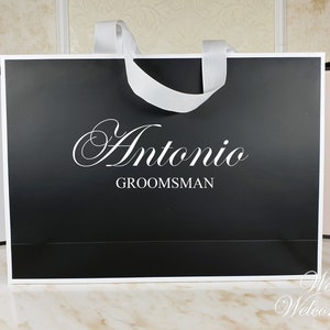 May include: Black paper gift bag with a white satin ribbon handle. The bag has the name "Antonio" and the word "Groomsman" printed in white on the front.