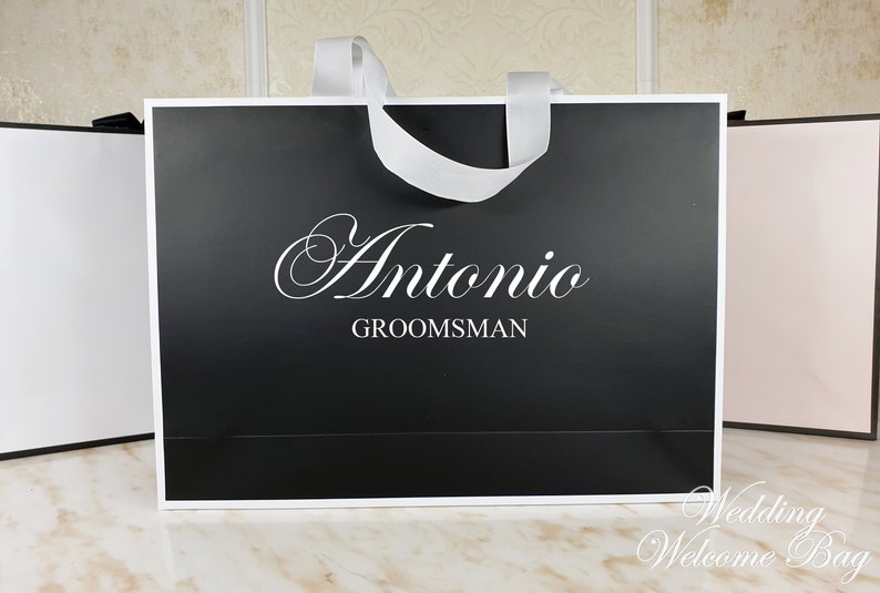 May include: Black paper gift bag with a white satin ribbon handle. The bag has the name "Antonio" and the word "Groomsman" printed in white on the front.