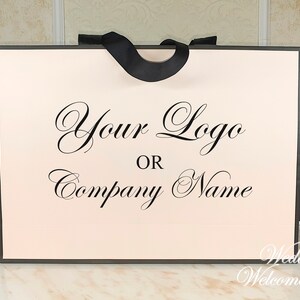 May include: A white wedding welcome bag with a black ribbon handle. The bag has a pink interior and the text "Your Logo or Company Name" printed in black cursive.