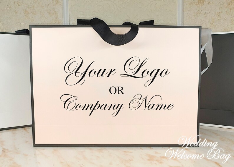 May include: A white wedding welcome bag with a black ribbon handle. The bag has a pink interior and the text "Your Logo or Company Name" printed in black cursive.
