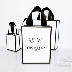 May include: Several white gift bags with black trim and handles are arranged on a white surface. The bags have the initials "E C" and the name "THOMPSON" with the date "22.02.22" printed on them. The bags are for a wedding.