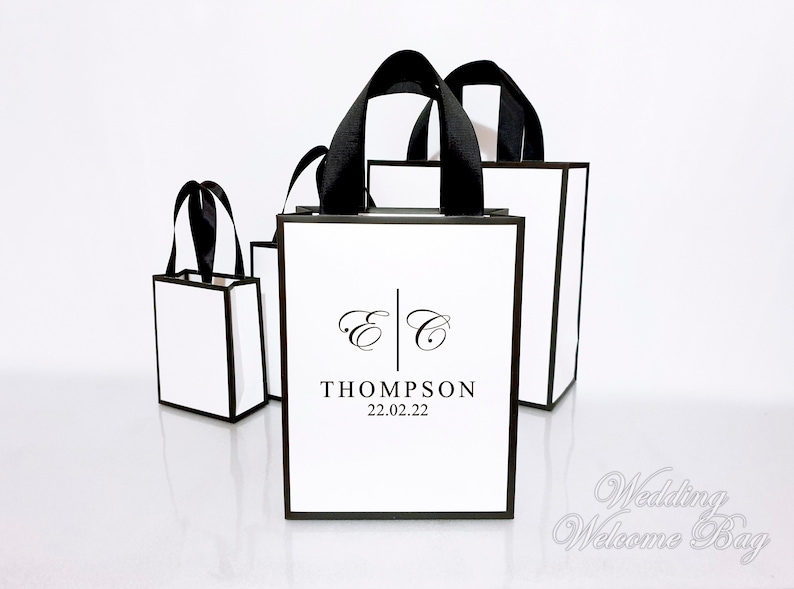 May include: Several white gift bags with black trim and handles are arranged on a white surface. The bags have the initials "E C" and the name "THOMPSON" with the date "22.02.22" printed on them. The bags are for a wedding.