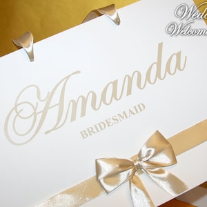 May include: White wedding welcome bag with a gold ribbon and bow. The bag is labeled "Amanda Bridesmaid".