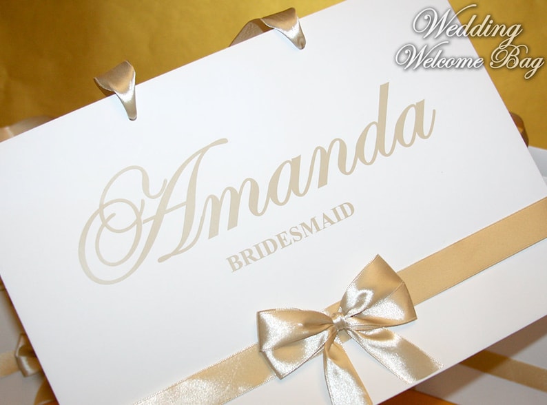 May include: White wedding welcome bag with a gold ribbon and bow. The bag is labeled "Amanda Bridesmaid".