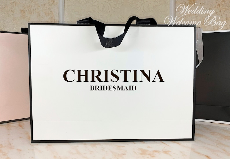 May include: White gift bag with black handles and a black border. The bag has the text "CHRISTINA BRIDESMAID" printed on the front.