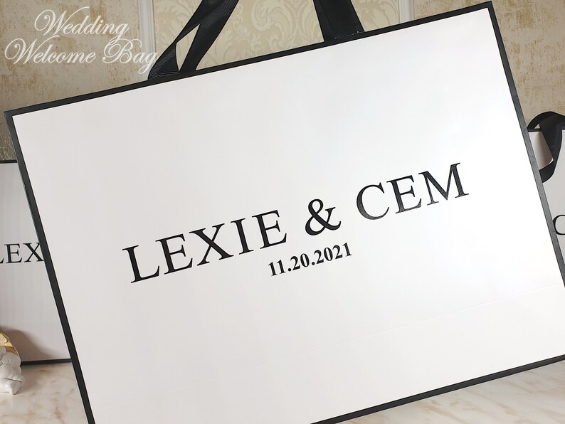 May include: White wedding welcome bag with black handles and black text that reads "Lexie & Cem 11.20.2021".