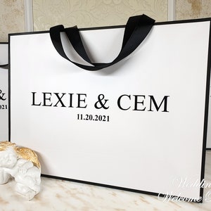 May include: White wedding welcome bag with black handles and the text "Lexie & Cem 11.20.2021" printed on the front.