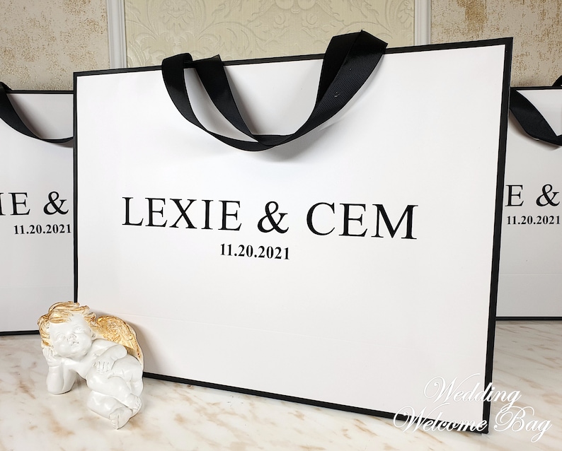 May include: White wedding welcome bag with black handles and the text "Lexie & Cem 11.20.2021" printed on the front.
