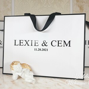 May include: Two white wedding welcome bags with black handles and the text "Lexie & Cem 11.20.2021" printed on the front. A small white angel figurine with gold wings is in front of the bag.
