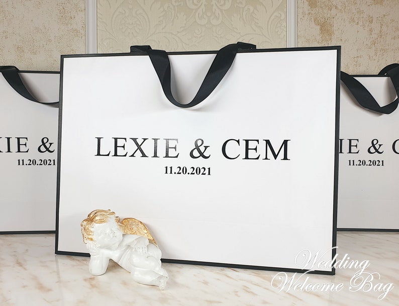 May include: Two white wedding welcome bags with black handles and the text "Lexie & Cem 11.20.2021" printed on the front. A small white angel figurine with gold wings is in front of the bag.