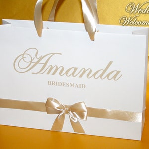 May include: White wedding gift bag with a gold ribbon and bow. The bag has the name "Amanda Bridesmaid" printed on it.