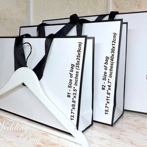 May include: Three white gift bags with black trim and handles. The bags are stacked on top of each other, with the largest bag on the bottom. The bags are labeled with the size of the bag in inches and centimeters. The text on the bags reads: "#1 - Size of bag 13.7"x9.8"x3.5" inches (35x25x9cm)", "#2 - Size of bag 15.7"x11.8"x4.7" inches (40x30x12cm)", and "#3 - Size of bag 19.6"x13.7"x5.9" inches (50x35x15cm)".