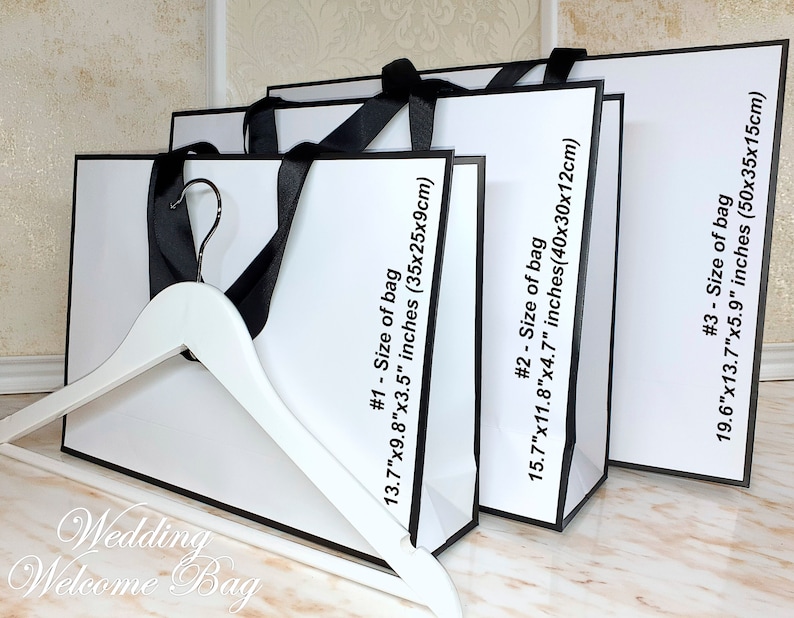 May include: Three white gift bags with black trim and handles. The bags are stacked on top of each other, with the largest bag on the bottom. The bags are labeled with the size of the bag in inches and centimeters. The text on the bags reads: "#1 - Size of bag 13.7"x9.8"x3.5" inches (35x25x9cm)", "#2 - Size of bag 15.7"x11.8"x4.7" inches (40x30x12cm)", and "#3 - Size of bag 19.6"x13.7"x5.9" inches (50x35x15cm)".