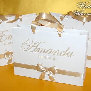 May include: Four white gift bags with gold ribbon and bows. The bags are personalized with the names "Emma Bride" and "Amanda Bridesmaid".