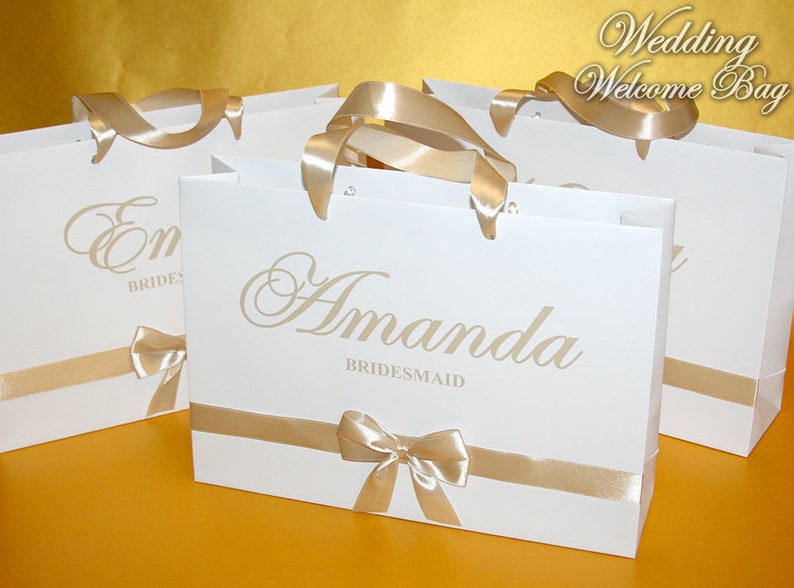 May include: Four white gift bags with gold ribbon and bows. The bags are personalized with the names "Emma Bride" and "Amanda Bridesmaid".