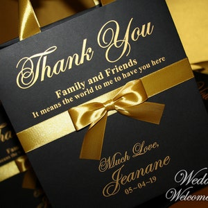 May include: Black wedding favor bag with a gold ribbon and bow. The bag has gold lettering that says "Thank You Family and Friends It means the world to me to have you here Much Love, Jeanane 05-04-19 Wedding Welcome Bag".