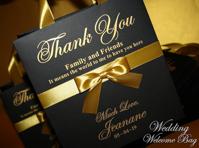 May include: Black wedding favor bag with a gold ribbon and bow. The bag has gold lettering that says "Thank You Family and Friends It means the world to me to have you here Much Love, Jeanane 05-04-19 Wedding Welcome Bag".