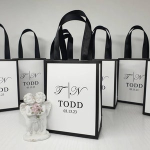 May include: White gift bags with black trim and handles are arranged in a row. Each bag features the initials "T | N" and the name "TODD" with the date "03.13.23" printed in black. A small white cherub statue holding flowers is in front of the bags.