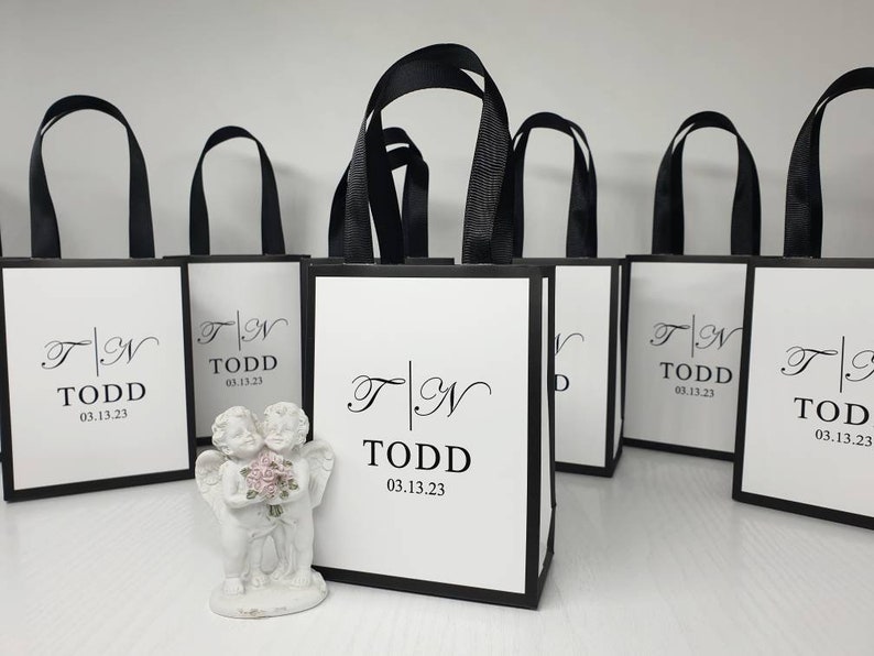 May include: White gift bags with black trim and handles are arranged in a row. Each bag features the initials "T | N" and the name "TODD" with the date "03.13.23" printed in black. A small white cherub statue holding flowers is in front of the bags.