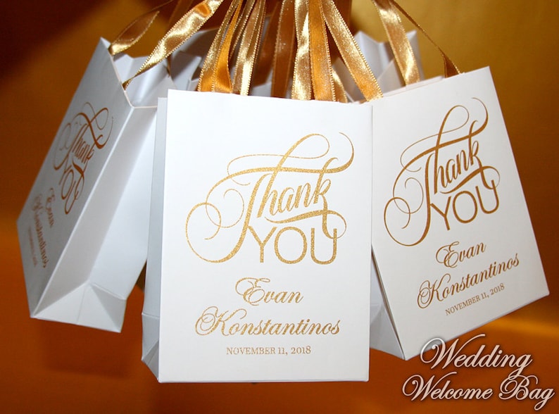Op de afbeelding: Witte bruiloft-goodiebags met gouden handvatten en gouden belettering met de tekst "Bedankt" en de namen "Evan Konstantinos" en de datum "11 november 2018".