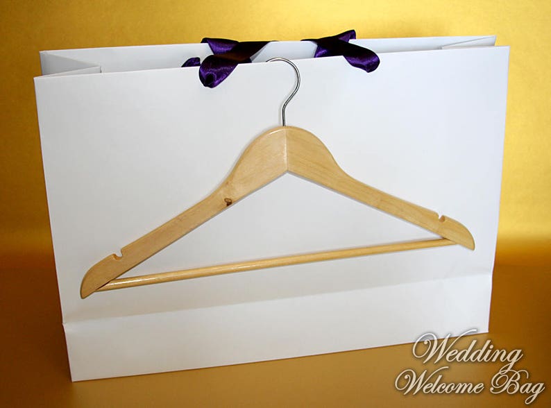 May include: White paper gift bag with purple ribbon handles and a wooden hanger inside. The bag has the text "Wedding Welcome Bag" printed on the bottom.