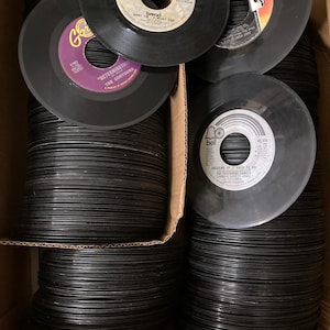 May include: A large collection of black vinyl 45 RPM records stacked in a cardboard box. The visible labels include Bread, The Contours, and The Partridge Family. Each record features a central hole and printed text detailing the artist and song.