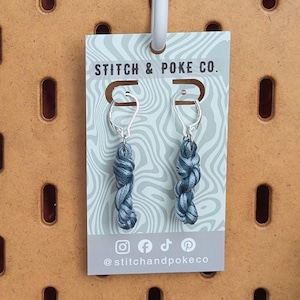 May include: A pair of silver-toned leverback earrings featuring small, twisted grey yarn skein charms. The earrings are displayed on a rectangular card labeled STITCH & POKE CO. with social media icons and the handle @stitchandpokeco printed at the bottom.