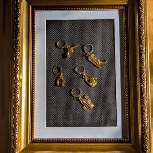 May include: Gold-colored earrings in a framed display. The earrings feature Western-themed designs: a gun, a cowboy boot, a horseshoe, a cowboy hat, and a cactus. The frame is ornate with a gold finish, set against a black background.