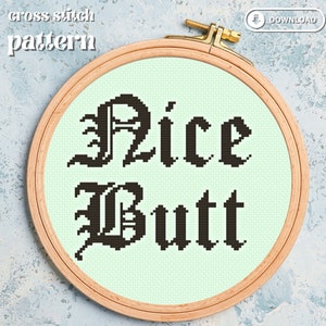 May include: A cross-stitch pattern in a wooden embroidery hoop. The design features the words "Nice Butt" in black, gothic-style lettering on a light green background. The words "cross stitch pattern" are above the hoop.