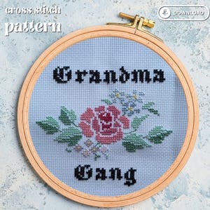 May include: A cross-stitch design in a wooden hoop displays the words "Grandma Gang" in black, gothic lettering. A pink rose with green leaves and blue flowers is centered on the light blue fabric.