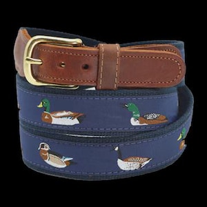 May include: A navy blue belt featuring an embroidered pattern of various ducks in shades of green, brown, and white. The belt includes a brown leather strap section with visible stitching and a polished gold-tone metal buckle with a single prong.